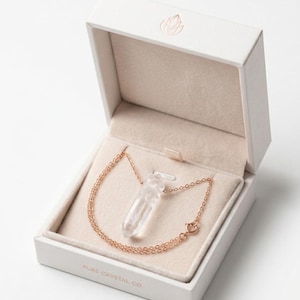 May include: A clear quartz crystal pendant necklace with a rose gold chain, presented in a white box with a soft beige interior. The box is labeled "PURE CRYSTAL CO."
