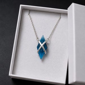 May include: A blue crystal pendant necklace with a silver chain and a silver X-shaped accent, presented in a white gift box. The crystal is diamond-shaped and has a faceted surface, reflecting light. The necklace is a piece of jewelry.