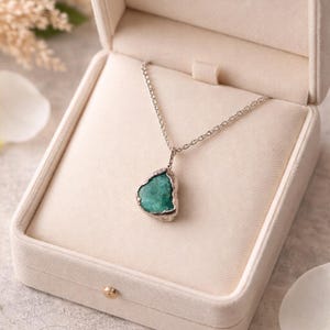Puede incluir: Collar de plata con un colgante de piedra preciosa de color turquesa. El colgante tiene forma tosca. El collar se presenta en un joyero crema abierto, con un interior suave y aterciopelado, sobre un fondo neutro.