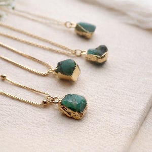 Puede incluir: Cuatro collares dorados con colgantes de piedras preciosas verde esmeralda. Los colgantes tienen una forma rugosa y natural y están engastados en oro. Los collares se exhiben sobre una superficie blanca, con una ramita de flores blancas en la esquina superior izquierda.