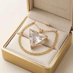 May include: A gold necklace with a large, clear, triangular pendant, presented in a gold jewelry box. The pendant is set in a gold frame and hangs from a delicate gold chain. The jewelry box is beige inside and has the text "SAGemsAndJewel" embossed on the lid.