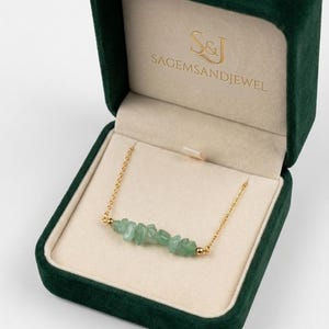 Peut inclure: Un collier à chaîne dorée orné d'une rangée de pierres vertes claires de forme irrégulière. Le collier est présenté dans un écrin à bijoux en velours vert foncé ouvert, avec le texte "S&J SAGEMSANDJEWEL" en lettres dorées.