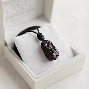 May include: A raw, deep red garnet gemstone pendant suspended from a thin black cord. The stone features a natural, irregular, and faceted texture. The necklace is presented inside an open, off-white, square-shaped jewelry box with a soft lining.