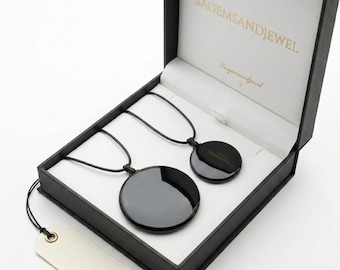 May include: Two black circular pendants on black cords inside a black box with a white interior. The box is open, revealing the pendants and the text "SAGEMSANDJEWEL" on the inside of the lid. A blank tag hangs from the box.