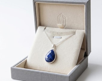 May include: A silver necklace with a teardrop-shaped, deep blue gemstone pendant, presented in a gray jewelry box with a light-colored interior. The box is open, showcasing the necklace's delicate chain and pendant.