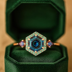 Puede incluir: Un anillo de oro con una gran gema azul hexagonal, rodeada de piedras verdes más pequeñas. Dos gemas triangulares púrpuras flanquean la piedra central. El anillo está en un joyero de terciopelo verde oscuro.