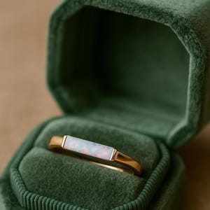 Könnte beinhalten: Ein goldener Ring mit einem rechteckigen Opalstein, der in einem goldenen Band gefasst ist. Der Ring wird in einer offenen, grünen Samt-Schmuckschatulle präsentiert. Der Opal hat eine weiße Basis mit Farbflecken.