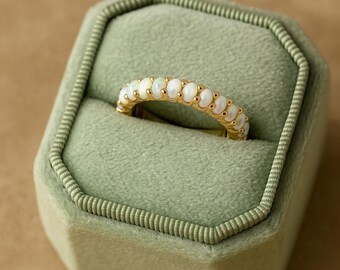 May include: A gold ring with a row of oval-shaped opals. The ring is set in a gold band and is displayed in a green velvet ring box. The opals are white with a slight iridescent sheen.