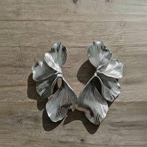 Puede incluir: Dos pendientes plateados con forma de hoja. Cada pendiente presenta tres hojas texturizadas en forma de abanico con vetas detalladas. Los pendientes están sobre una superficie de madera.