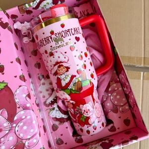 May include: A pink Stanley tumbler with a red handle and lid, featuring a Strawberry Shortcake design. The tumbler is decorated with strawberries, hearts, and a cartoon character. It comes in a matching pink box with strawberry and polka dot patterns.