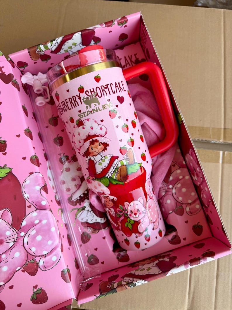 May include: A pink Stanley tumbler with a red handle and lid, featuring a Strawberry Shortcake design. The tumbler is decorated with strawberries, hearts, and a cartoon character. It comes in a matching pink box with strawberry and polka dot patterns.