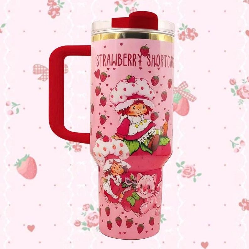 May include: Pink insulated tumbler with a red handle and lid. The tumbler features a Strawberry Shortcake design with illustrations of the character, strawberries, and the text "Strawberry Shortcake".