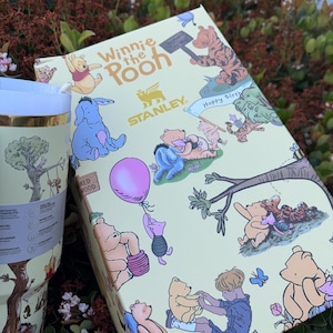 Op de afbeelding: Een Winnie de Poeh-thema cadeau set met een beker en een doos. De doos en de beker hebben een lichtgele achtergrond met illustraties van Winnie de Poeh en zijn vrienden. De beker heeft een gouden rand en een boomillustratie.