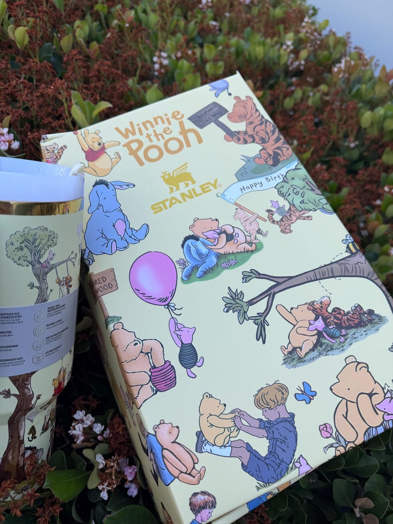 Op de afbeelding: Een Winnie de Poeh-thema cadeau set met een beker en een doos. De doos en de beker hebben een lichtgele achtergrond met illustraties van Winnie de Poeh en zijn vrienden. De beker heeft een gouden rand en een boomillustratie.
