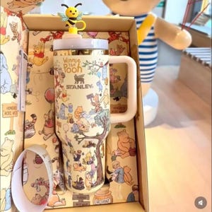 Może przedstawiać: Kremowy kubek Stanley z postaciami Kubusia Puchatka i napisem "Winnie the Pooh". Kubek ma biały uchwyt i ż&oacute;łtą nakładkę w kształcie pszczoły. Kubek znajduje się w pudełku z pasującymi ilustracjami.