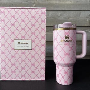 May include: A pink Stanley Loveshackfancy tumbler with a handle and a matching box. Both the tumbler and box feature a pink bow pattern. The tumbler has a clear lid and gold accents. The Stanley and Loveshackfancy logos are visible.