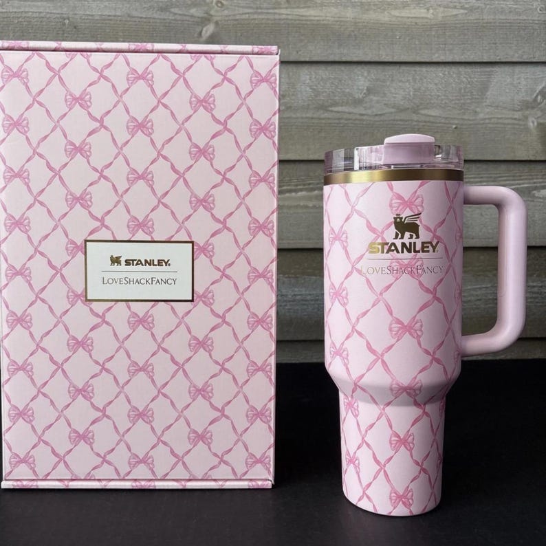 May include: A pink Stanley Loveshackfancy tumbler with a handle and a matching box. Both the tumbler and box feature a pink bow pattern. The tumbler has a clear lid and gold accents. The Stanley and Loveshackfancy logos are visible.