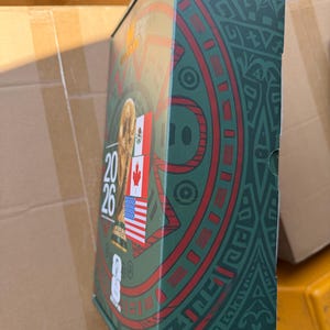 Op de afbeelding: Een rechthoekige FIFA World Cup 2026 herdenkingsblik met een groene achtergrond en ingewikkelde donkergroene patronen. De voorzijde toont de FIFA World Cup trofee, de tekst 2026 en de vlaggen van Mexico, Canada en de Verenigde Staten.