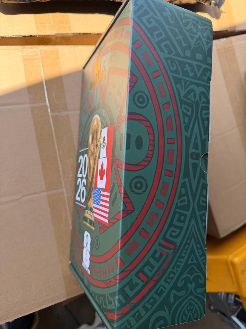 Op de afbeelding: Een rechthoekige FIFA World Cup 2026 herdenkingsblik met een groene achtergrond en ingewikkelde donkergroene patronen. De voorzijde toont de FIFA World Cup trofee, de tekst 2026 en de vlaggen van Mexico, Canada en de Verenigde Staten.