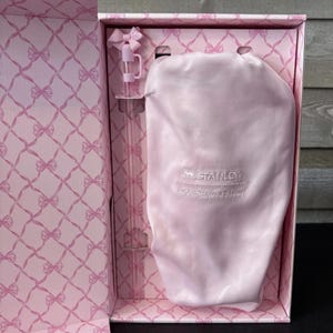 May include: A pink gift box with a bow-shaped lid and a matching pink interior pattern. Inside, a pink Stanley Loveshackfancy water bottle and a pink satin drawstring bag are visible.