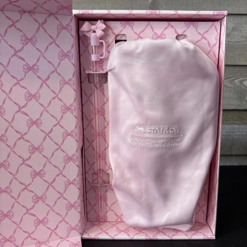 May include: A pink gift box with a bow-shaped lid and a matching pink interior pattern. Inside, a pink Stanley Loveshackfancy water bottle and a pink satin drawstring bag are visible.