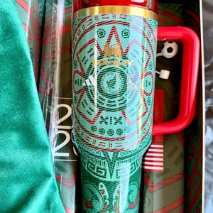 May include: A green and red Stanley travel mug with a geometric design and a red handle and lid. The mug has a gold rim and a small soccer ball charm attached to the handle. The mug is in a green box.