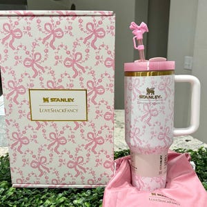 K&ouml;nnte beinhalten: Ein wei&szlig;-rosa Stanley LoveShackFancy Becher mit floralem Muster, Henkel und Strohhalm. Der Becher befindet sich in einer passenden Box mit dem gleichen floralen Muster. Die Box und der Becher haben die Logos von Stanley und LoveShackFancy.