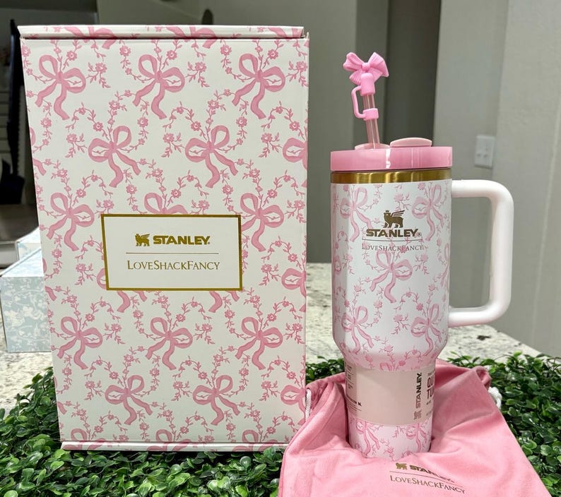 K&ouml;nnte beinhalten: Ein wei&szlig;-rosa Stanley LoveShackFancy Becher mit floralem Muster, Henkel und Strohhalm. Der Becher befindet sich in einer passenden Box mit dem gleichen floralen Muster. Die Box und der Becher haben die Logos von Stanley und LoveShackFancy.