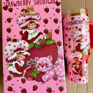 May include: A pink Strawberry Shortcake themed gift set featuring a box and a tumbler. The box has illustrations of Strawberry Shortcake and a pink cat. The tumbler has a red handle and lid, with the words "Strawberry Shortcake" printed on it.