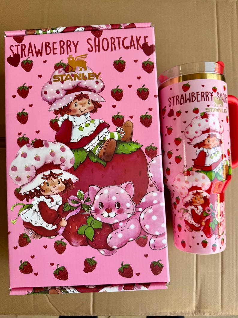 May include: A pink Strawberry Shortcake themed gift set featuring a box and a tumbler. The box has illustrations of Strawberry Shortcake and a pink cat. The tumbler has a red handle and lid, with the words "Strawberry Shortcake" printed on it.