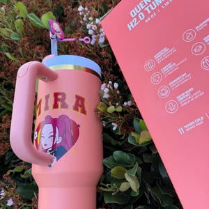 Puede incluir: Un vaso Quencher H2.0 rosa de 1,18 L con asa, con un gr&aacute;fico de dibujos animados de una mujer con pelo rosa y el nombre "MIRA". El vaso tiene una pajita y una tapa. El embalaje tambi&eacute;n es rosa.