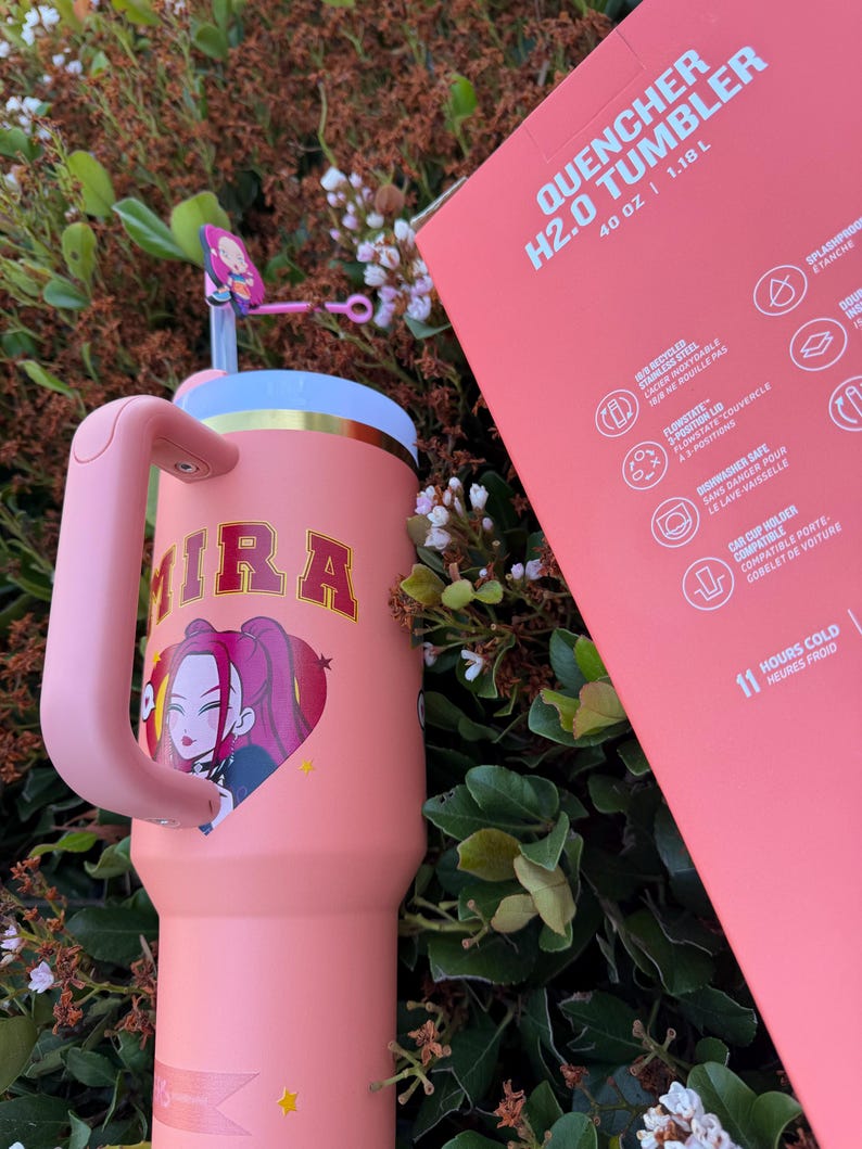 Puede incluir: Un vaso Quencher H2.0 rosa de 1,18 L con asa, con un gr&aacute;fico de dibujos animados de una mujer con pelo rosa y el nombre "MIRA". El vaso tiene una pajita y una tapa. El embalaje tambi&eacute;n es rosa.