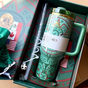 Op de afbeelding: Een groene Stanley Quencher H2.0 beker met een patroon, een bijpassend groen handvat en een doorzichtig rietje met een kleine voetbalbedel. De beker zit in een decoratieve doos met vlaggen en een goudkleurige afbeelding van een schedel.
