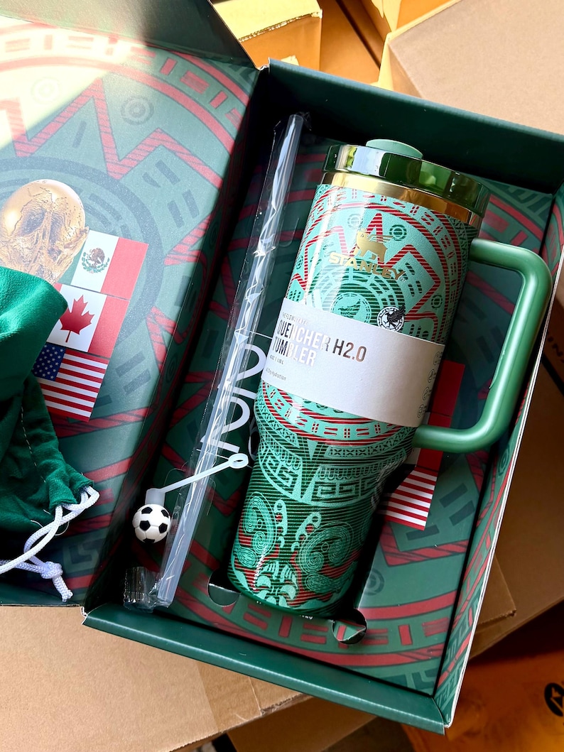Op de afbeelding: Een groene Stanley Quencher H2.0 beker met een patroon, een bijpassend groen handvat en een doorzichtig rietje met een kleine voetbalbedel. De beker zit in een decoratieve doos met vlaggen en een goudkleurige afbeelding van een schedel.