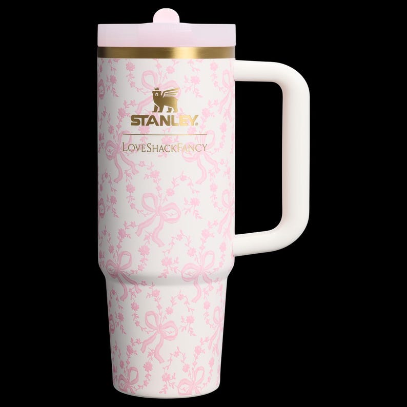 K&ouml;nnte beinhalten: Ein wei&szlig;-rosa Stanley-Becher mit floralem Muster und Griff. Der Becher hat einen rosa Deckel und ein goldenes Band. Der Text "STANLEY" und "LOVESHACKFANCY" sind auf dem Becher aufgedruckt.