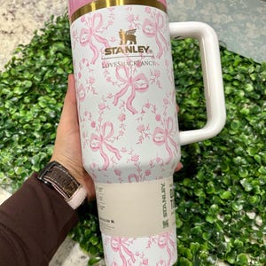 K&ouml;nnte beinhalten: Ein gro&szlig;er, wei&szlig;er Stanley-Becher mit rosa Deckel und Griff, verziert mit einem rosa Schleifen- und Blumenmuster. Der Becher tr&auml;gt das Logo "LOVESHACKFANCY" und ein goldenes Band. Der Griff ist wei&szlig;.