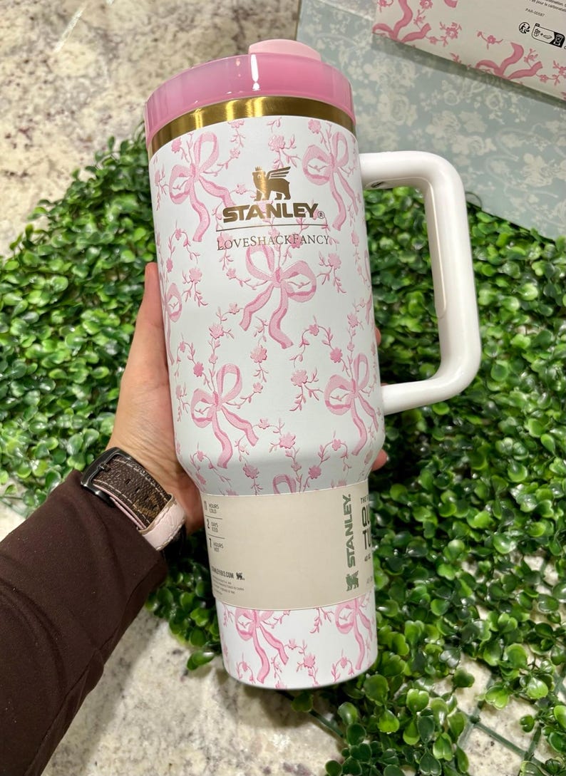 K&ouml;nnte beinhalten: Ein gro&szlig;er, wei&szlig;er Stanley-Becher mit rosa Deckel und Griff, verziert mit einem rosa Schleifen- und Blumenmuster. Der Becher tr&auml;gt das Logo "LOVESHACKFANCY" und ein goldenes Band. Der Griff ist wei&szlig;.