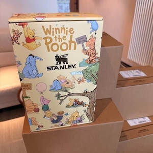 Może przedstawiać: Ż&oacute;łte pudełko Stanley z postaciami Kubusia Puchatka. Pudełko zawiera ilustracje Kubusia Puchatka, Tygryska, Kłapouchego i Prosiaczka. Pudełko stoi na kilku brązowych kartonach.