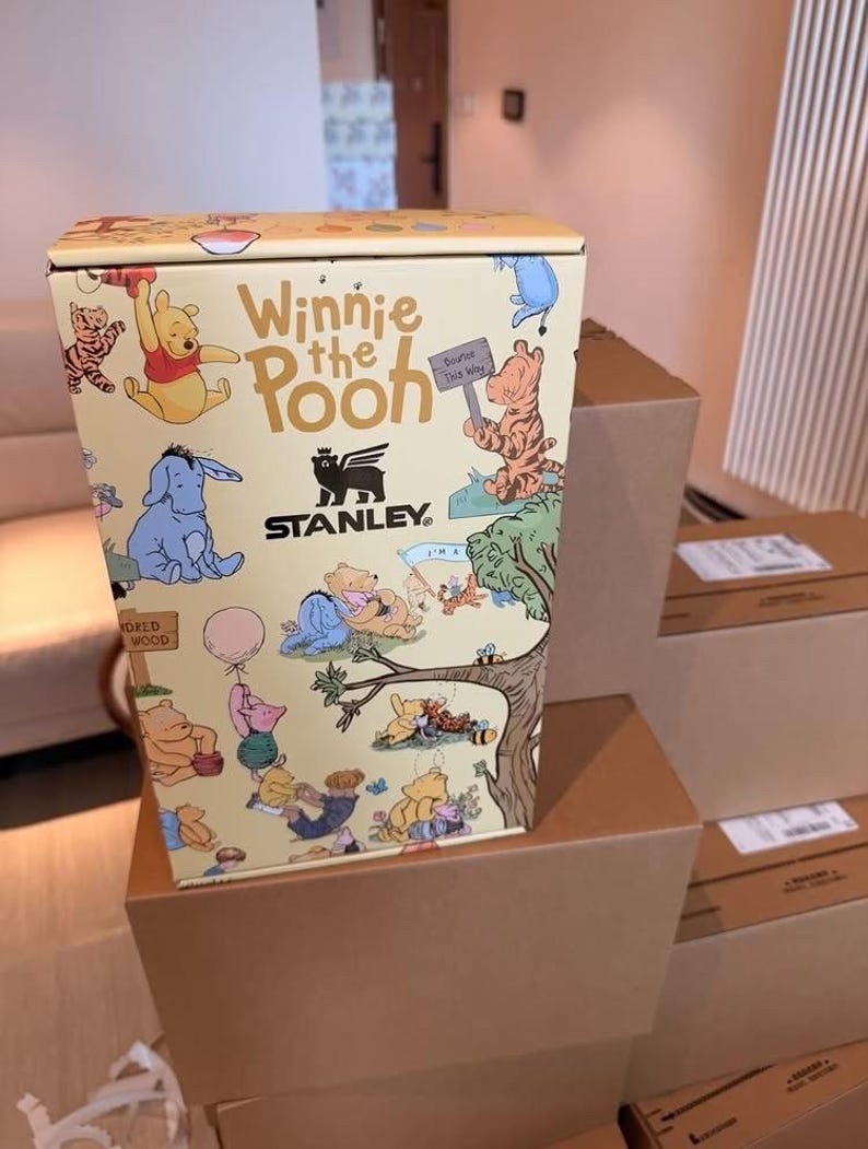 Może przedstawiać: Ż&oacute;łte pudełko Stanley z postaciami Kubusia Puchatka. Pudełko zawiera ilustracje Kubusia Puchatka, Tygryska, Kłapouchego i Prosiaczka. Pudełko stoi na kilku brązowych kartonach.