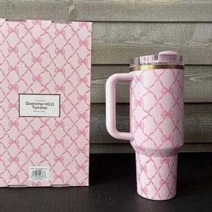 May include: A pink tumbler with a handle and a clear lid, featuring a repeating pattern of bows. The tumbler is next to a matching box with the text "Quencher H2.0 Tumbler" and a capacity of 40 oz (1.18 L).