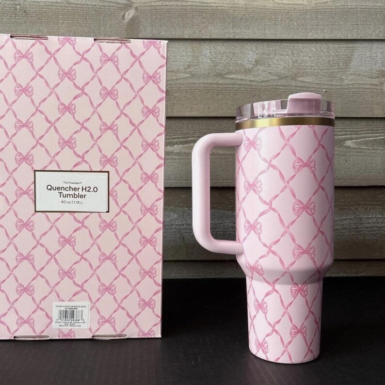 May include: A pink tumbler with a handle and a clear lid, featuring a repeating pattern of bows. The tumbler is next to a matching box with the text "Quencher H2.0 Tumbler" and a capacity of 40 oz (1.18 L).