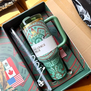 Op de afbeelding: Een groene Stanley Quencher H2.0 beker met een rood en groen patroon, een handvat en een doorzichtig rietje met een kleine voetbalbedel. De beker zit in een decoratieve doos met de vlaggen van Mexico, Canada en de Verenigde Staten.