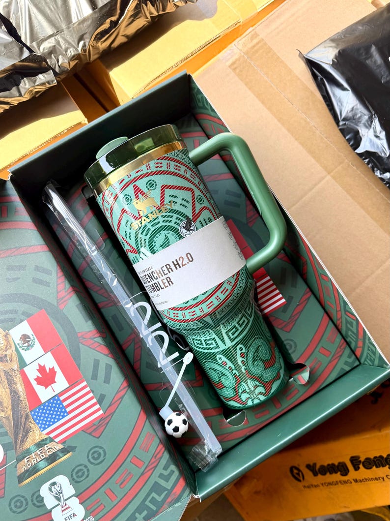 Op de afbeelding: Een groene Stanley Quencher H2.0 beker met een rood en groen patroon, een handvat en een doorzichtig rietje met een kleine voetbalbedel. De beker zit in een decoratieve doos met de vlaggen van Mexico, Canada en de Verenigde Staten.