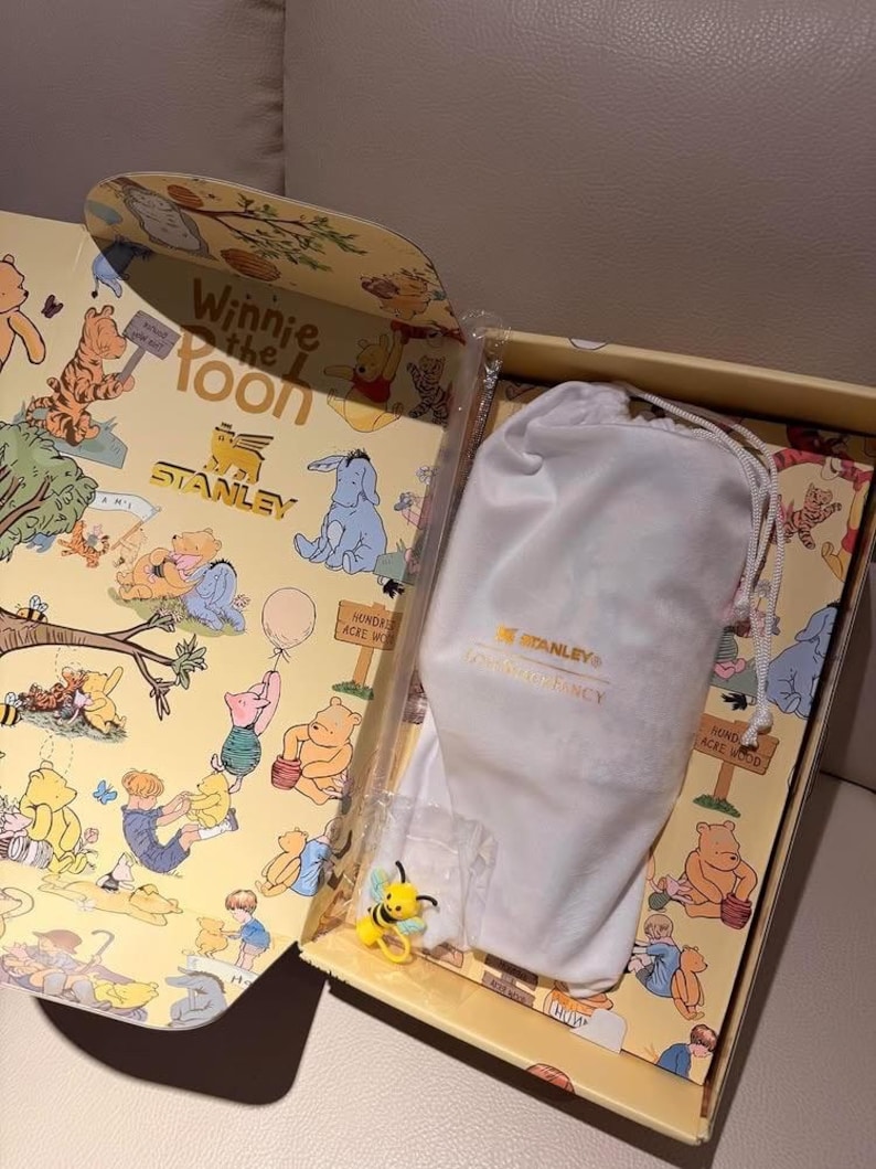 Może przedstawiać: Pudełko prezentowe z motywem Kubusia Puchatka, z ilustracjami Kubusia, Prosiaczka, Tygryska i Kłapouchego. Zawiera biały worek ze sznurkiem z logo Stanley i ż&oacute;łto-czarną figurkę pszczoły.