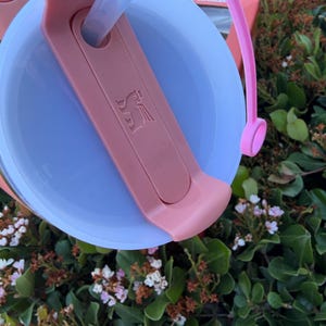 Puede incluir: Primer plano de una tapa de vaso rosa y azul con pajita. La tapa tiene un asa rosa y un deslizador rosa con un logotipo. El vaso est&aacute; parcialmente lleno de un l&iacute;quido transparente. El fondo es follaje verde.