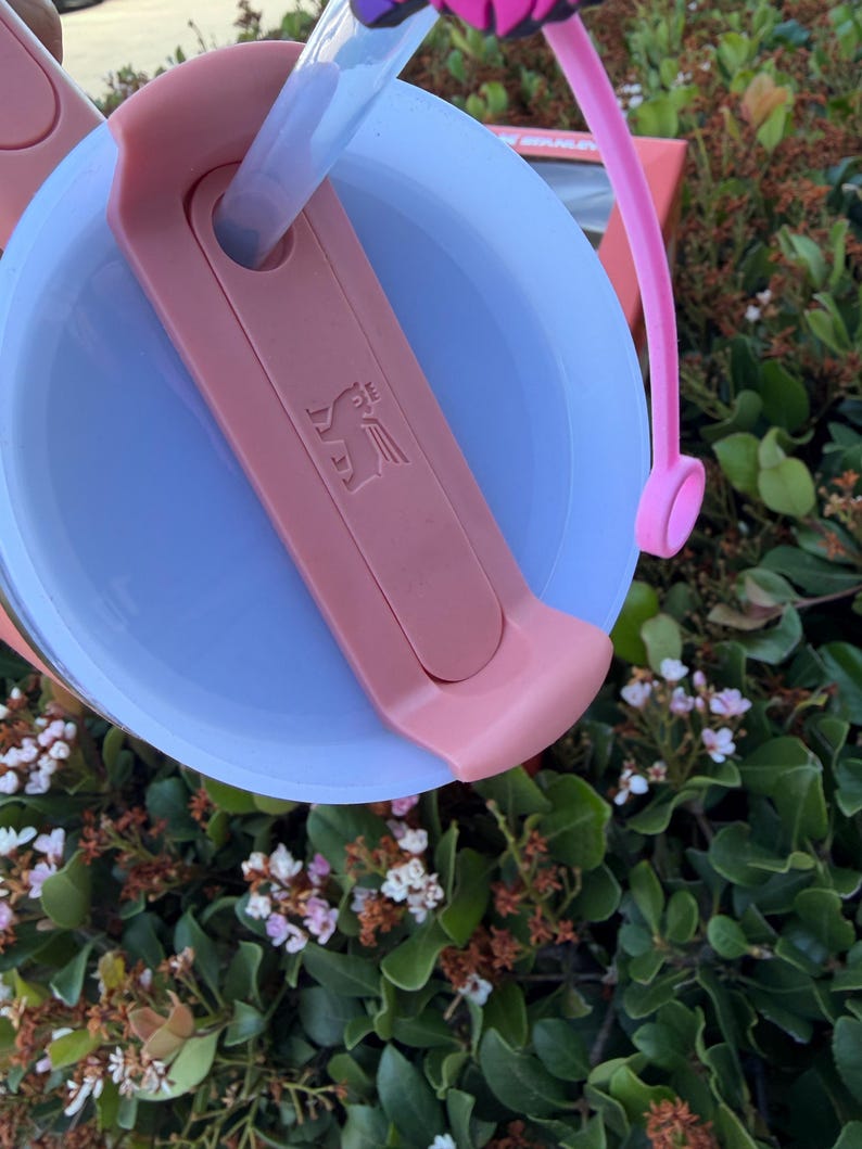 Puede incluir: Primer plano de una tapa de vaso rosa y azul con pajita. La tapa tiene un asa rosa y un deslizador rosa con un logotipo. El vaso est&aacute; parcialmente lleno de un l&iacute;quido transparente. El fondo es follaje verde.