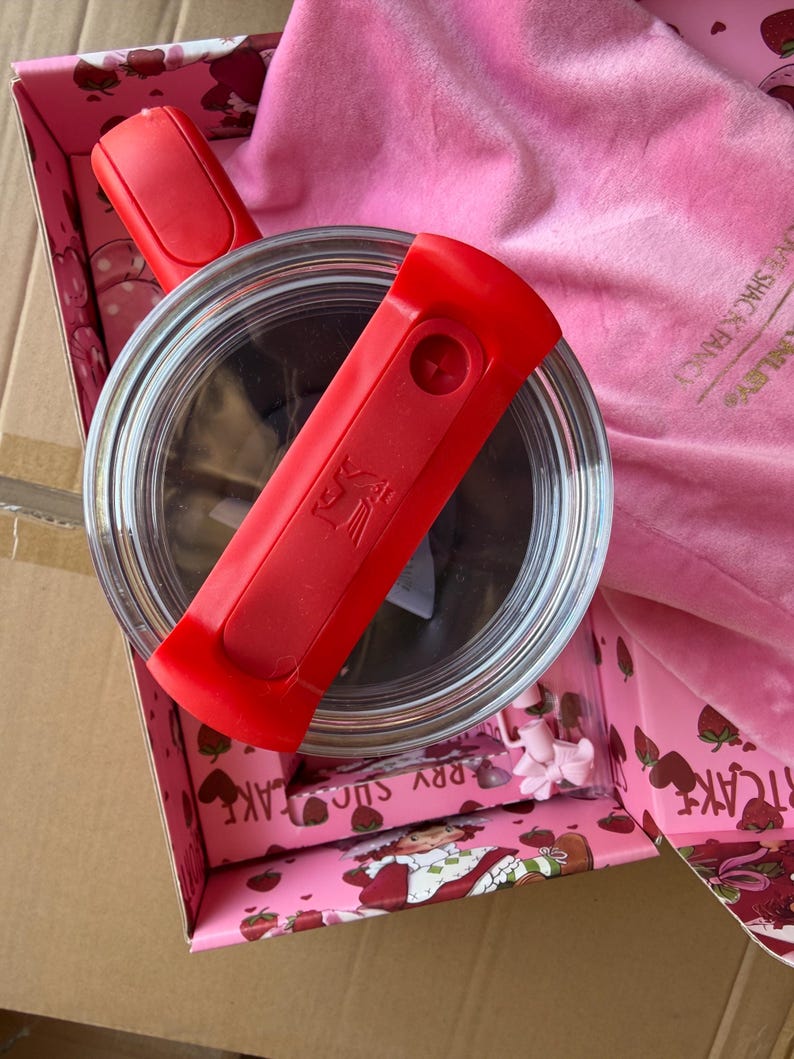 May include: A clear glass container with a red handle and a red lid is inside a pink box with a strawberry design. A pink cloth and small pink accessories are also in the box. The box is inside a larger cardboard box.