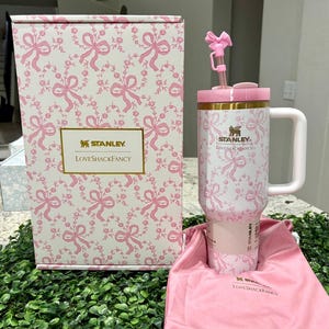 K&ouml;nnte beinhalten: Ein wei&szlig;-rosa Stanley Loveshackfancy Becher mit Griff, Strohhalm und Schleifendetail. Der Becher und die Box haben ein rosa Schleifen- und Blumenmuster. Ein rosa Kordelzugbeutel ist ebenfalls enthalten.