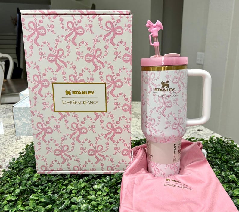 K&ouml;nnte beinhalten: Ein wei&szlig;-rosa Stanley Loveshackfancy Becher mit Griff, Strohhalm und Schleifendetail. Der Becher und die Box haben ein rosa Schleifen- und Blumenmuster. Ein rosa Kordelzugbeutel ist ebenfalls enthalten.