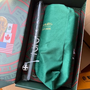 Op de afbeelding: Een groene stoffen hoes met de gouden tekst STANLEY CUP 2026, een doorzichtig plastic rietje met een kleine voetbalbedel en een groene doos met de vlaggen van Mexico, Canada en de Verenigde Staten op de binnenzijde van het deksel.